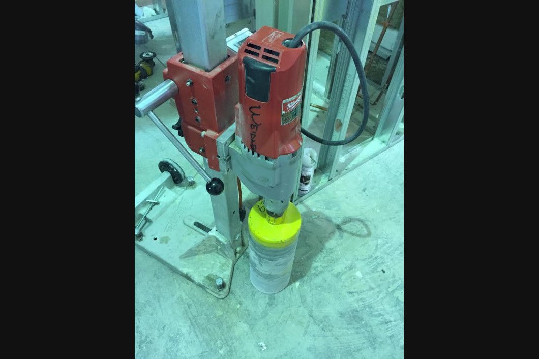 A red core drill is drilling a hole into concrete, with a yellow drill bit and a gray base.