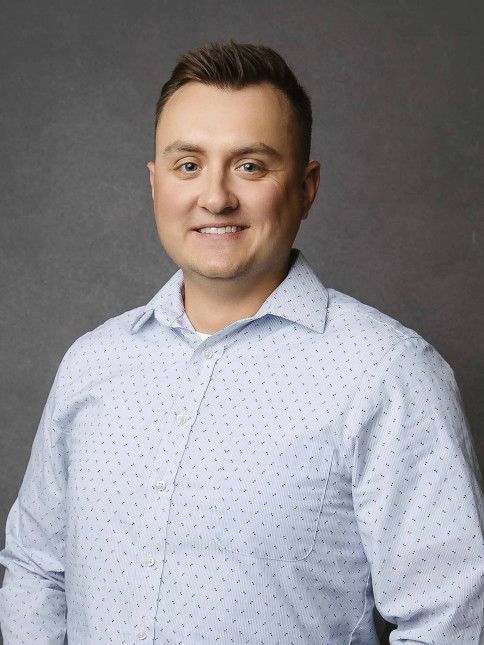 Man wearing light blue button-down shirt smiles in front of gray background.