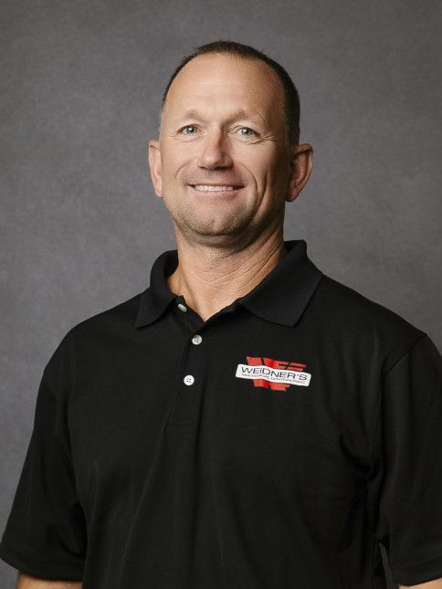 Man in black polo shirt smiling at camera. Gray background.