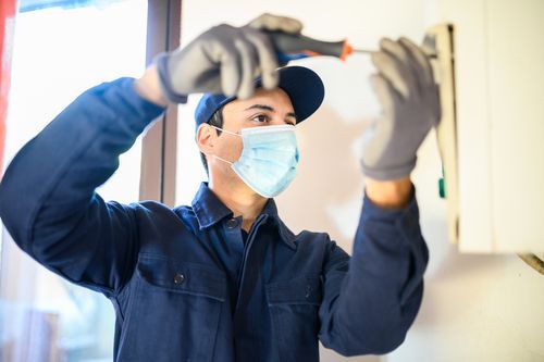Person wearing gloves and a face mask uses a screwdriver on a wall-mounted appliance.