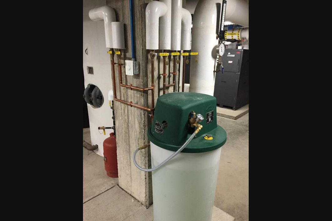 Water softener in basement near pipes and furnace. Green top, white tank, copper piping.