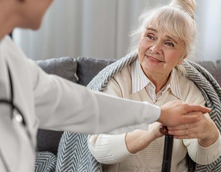 Caregiver holding hands with a smiling elderly woman on a couch, with a blanket around her shoulders.