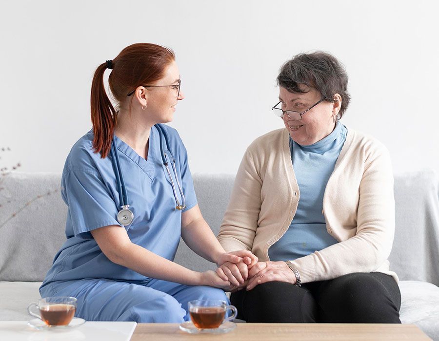 Nurse in blue scrubs holding hands with a seated patient in a bright clinic room, with tea cups on the table