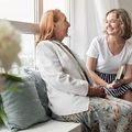 Two women chatting on a couch in a bright living room, smiling and holding books.