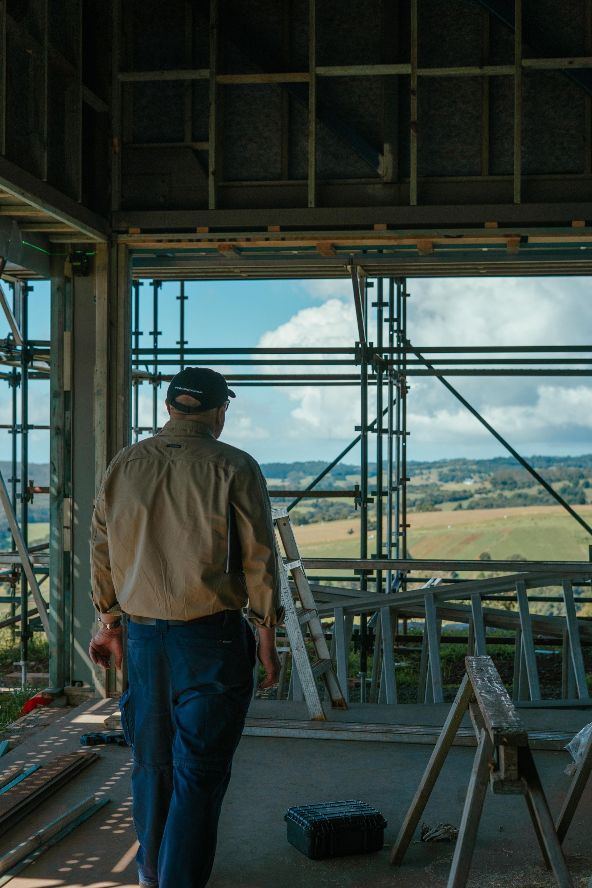 Worker standing in unfinished building, looking out over scaffolding and fields through large open frame.