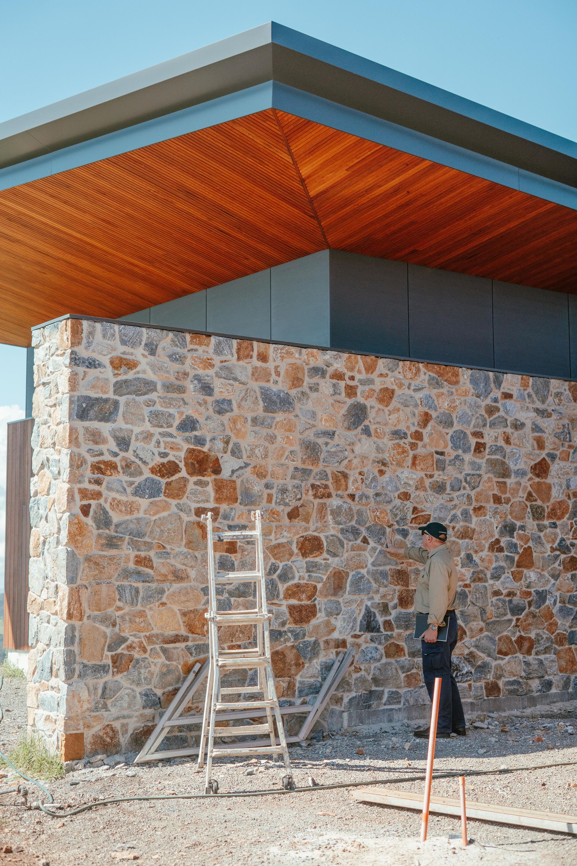 Stone building exterior with wood overhang, ladder, and worker on gravel site