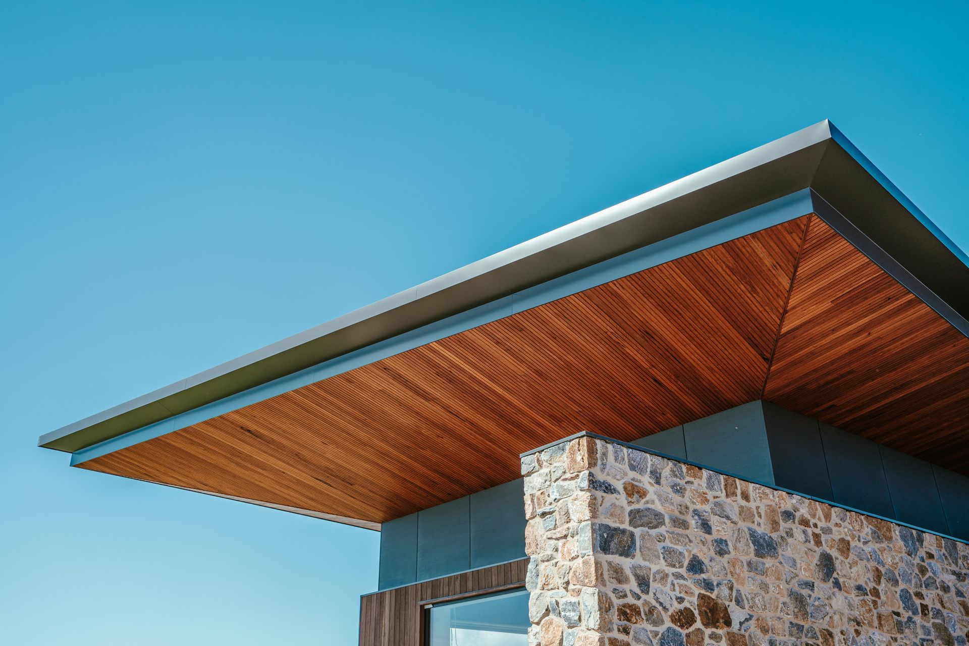 Modern building corner with wood soffit and stone wall under a blue sky