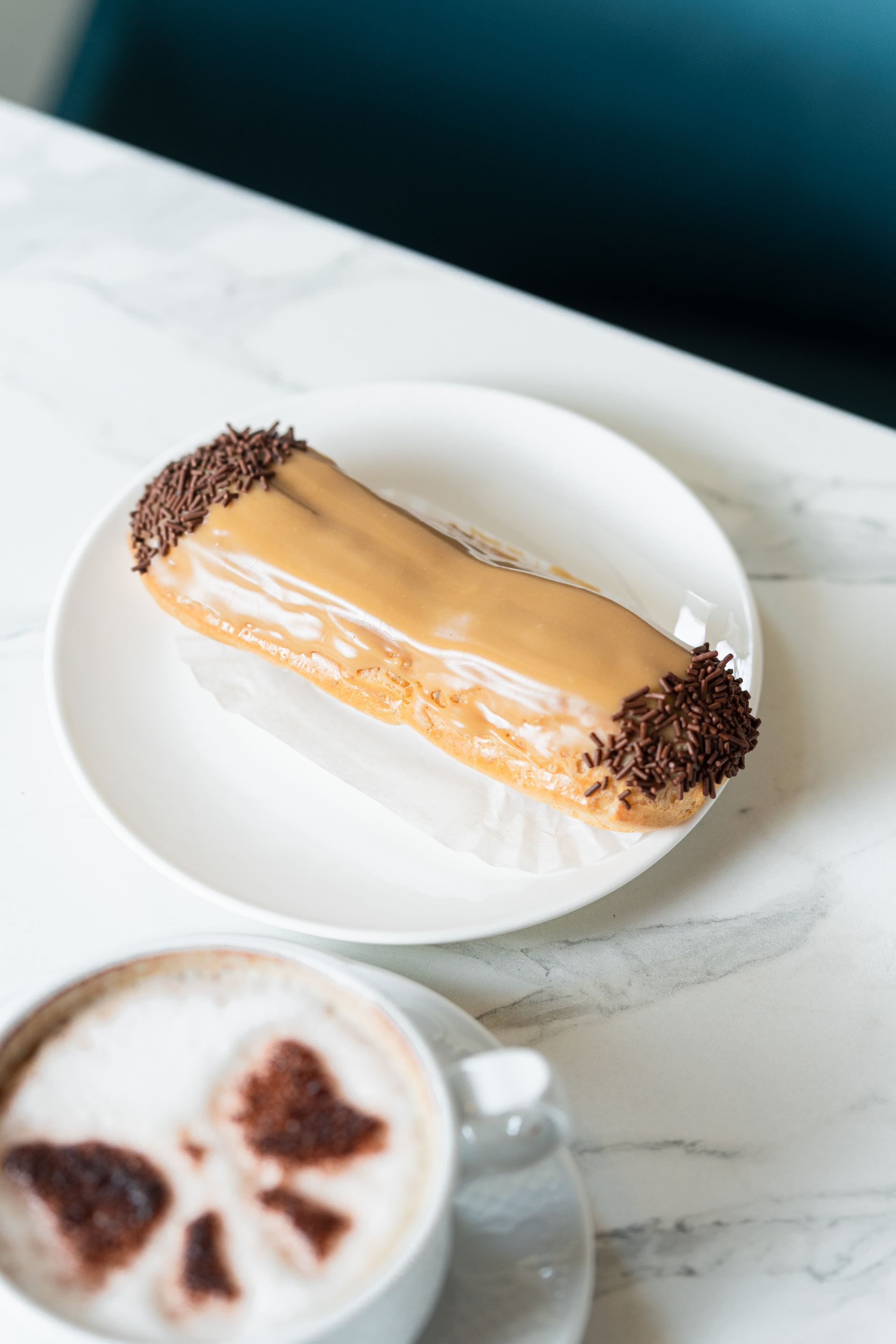 Pâtisserie fourrée à la crème et nappée de chocolat, présentée sur une assiette blanche à côté d'un cappuccino sur une table en marbre.