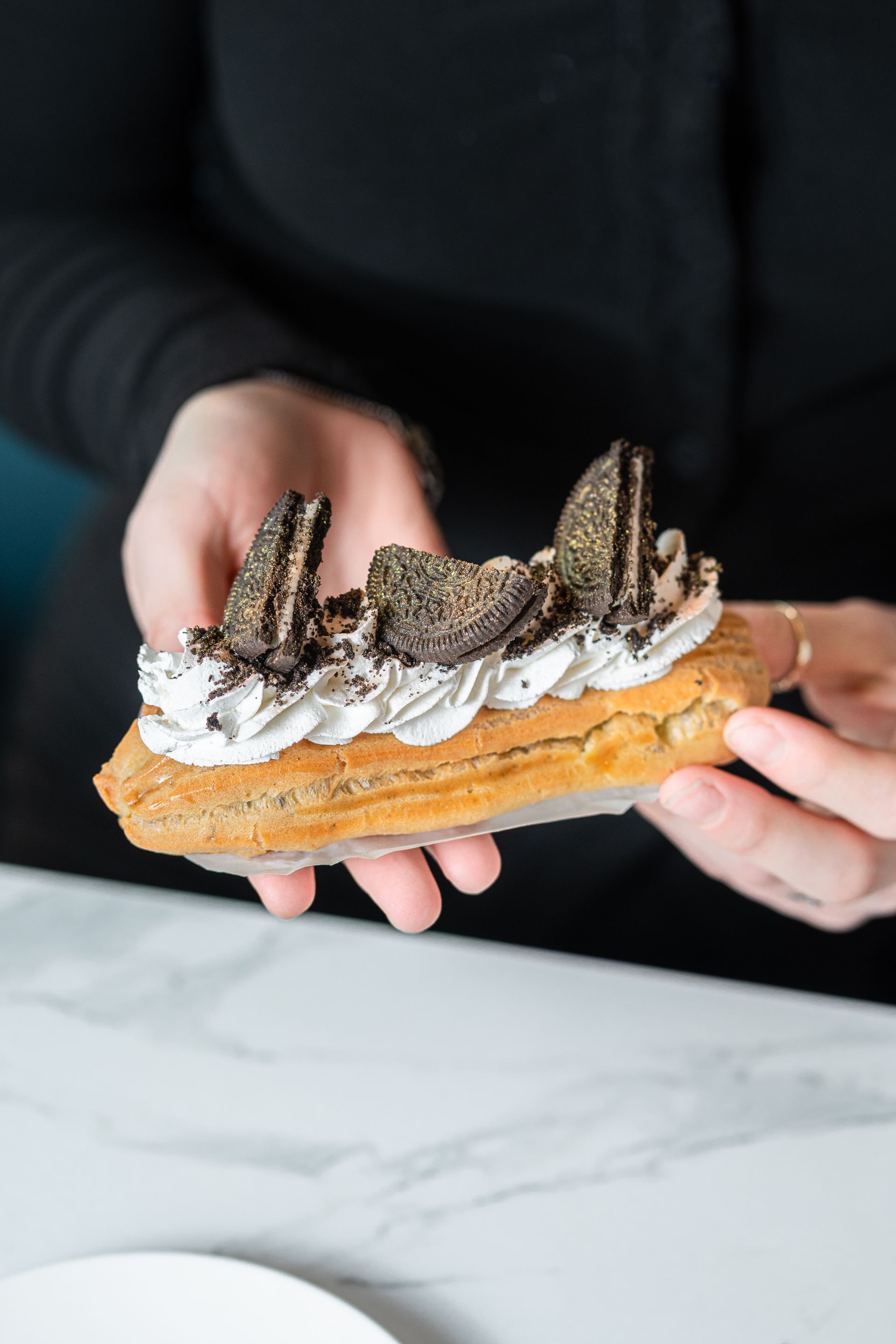 Des mains tenant une longue pâtisserie garnie de crème fouettée, de décorations en chocolat et de morceaux de biscuits, posée sur un comptoir en marbre.