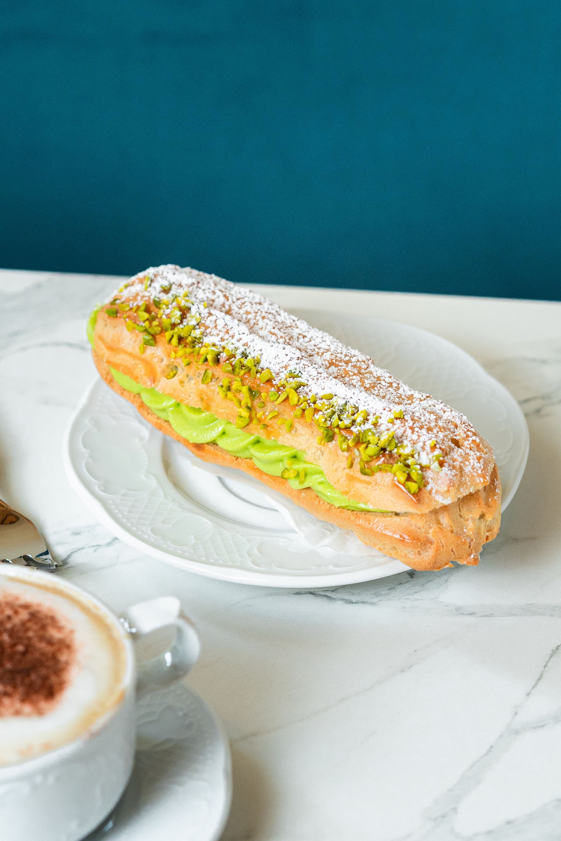 Éclair à la pistache en poudre sur une assiette blanche à côté d'une tasse de café