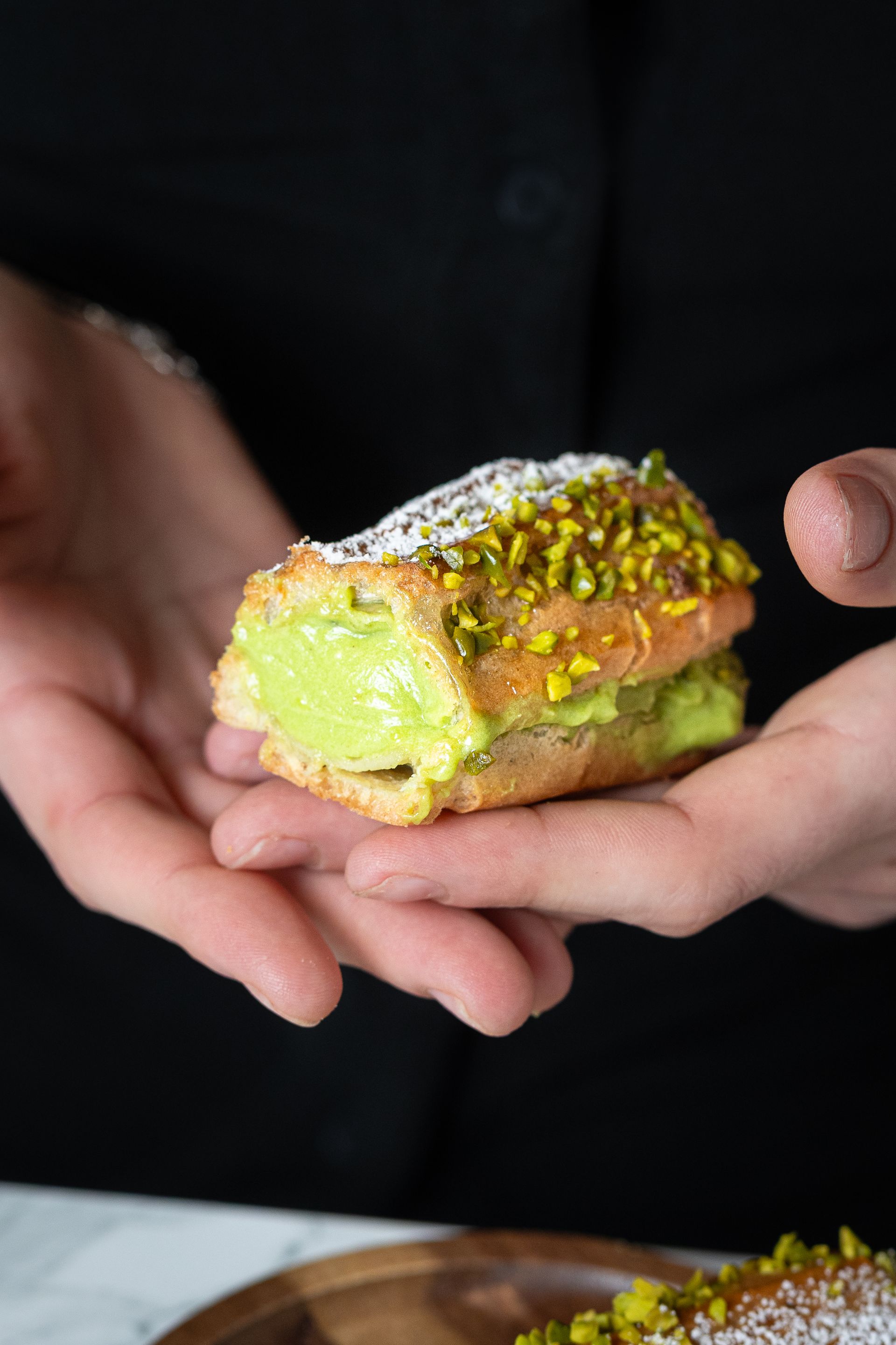 Des mains tenant un carré de dessert enrobé de pistaches et fourré d'une crème onctueuse.