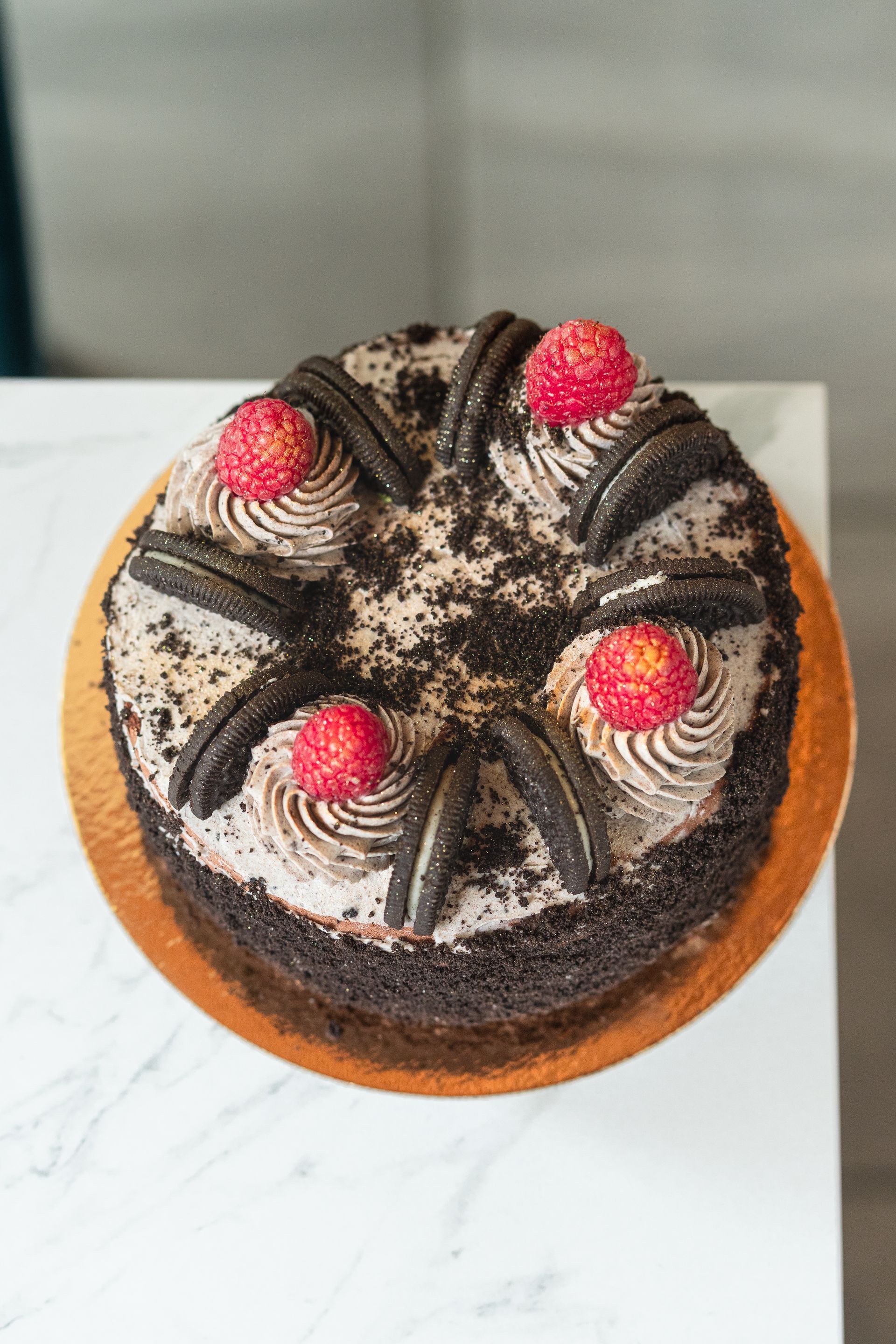 Gâteau au chocolat garni de volutes de crème, de miettes d'Oreo et de framboises rouges, présenté sur une planche blanche.