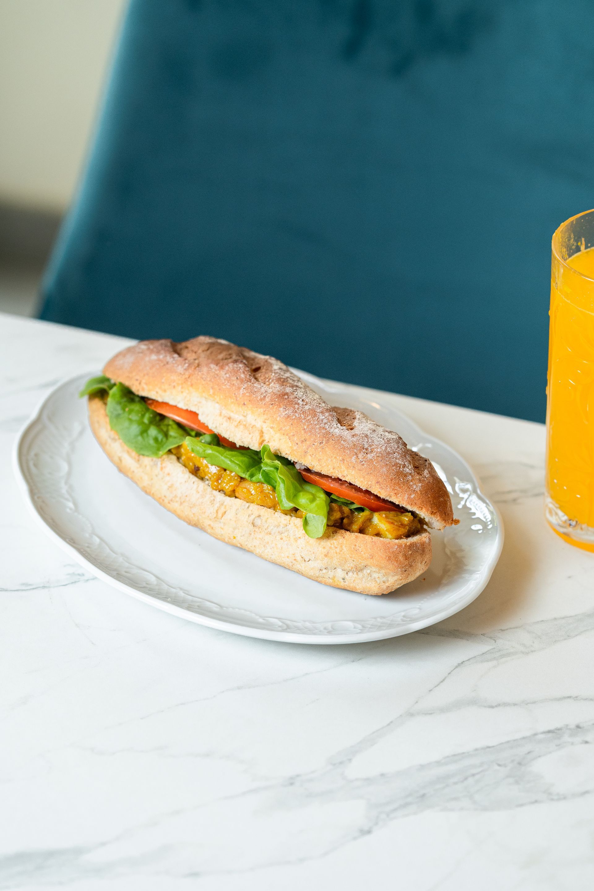 Un sandwich sur une assiette blanche à côté d'un verre de jus d'orange sur une table en marbre