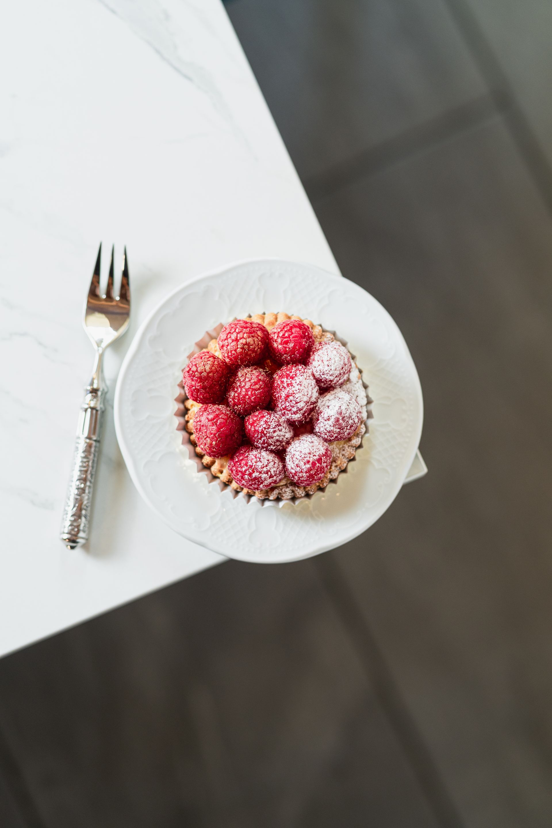 Un bol de framboises saupoudrées de sucre glace à côté d'une fourchette sur une table blanche
