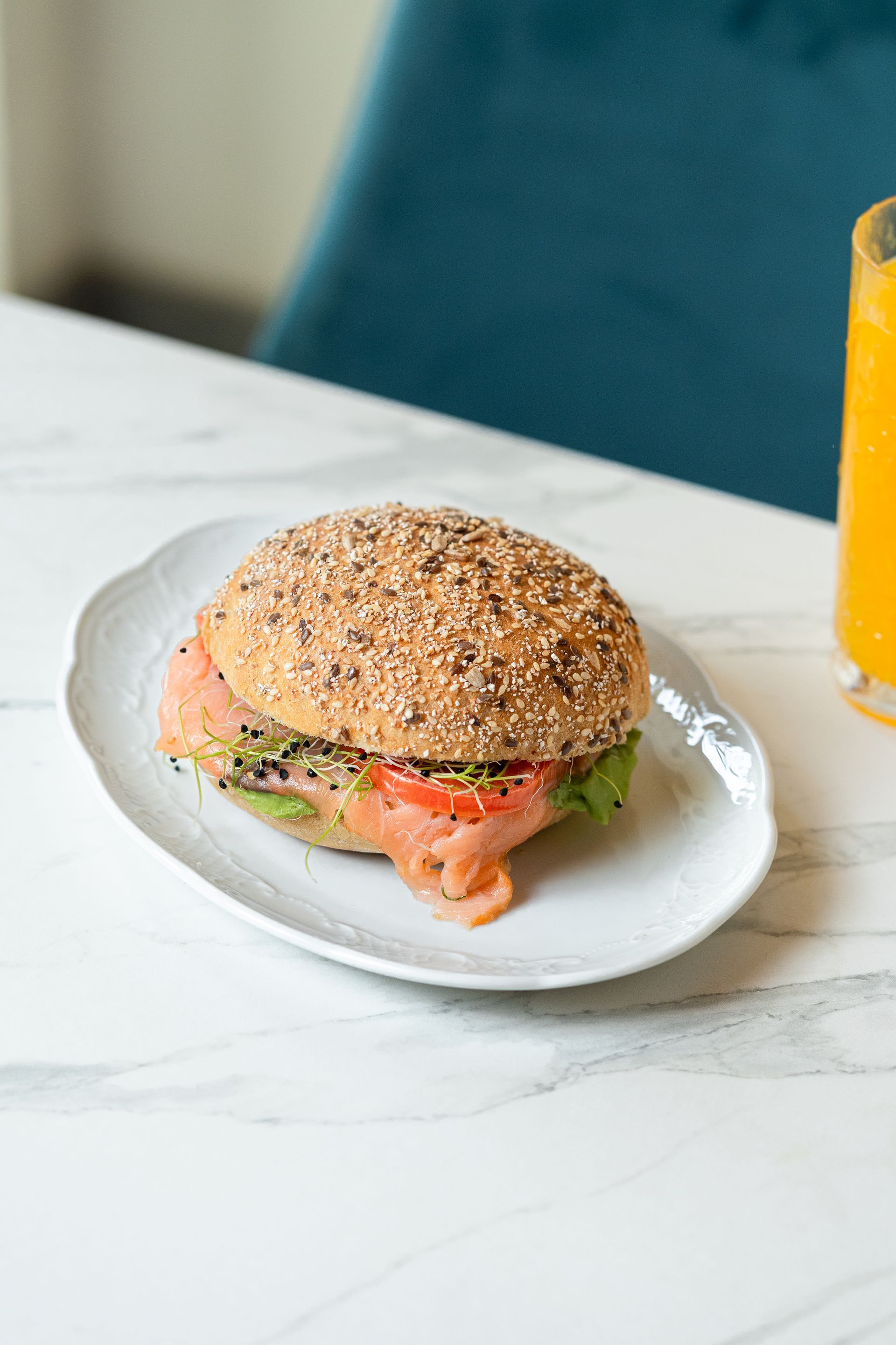 Un sandwich aux graines de sésame sur une assiette blanche, à côté d'un verre de jus d'orange sur une table en marbre.