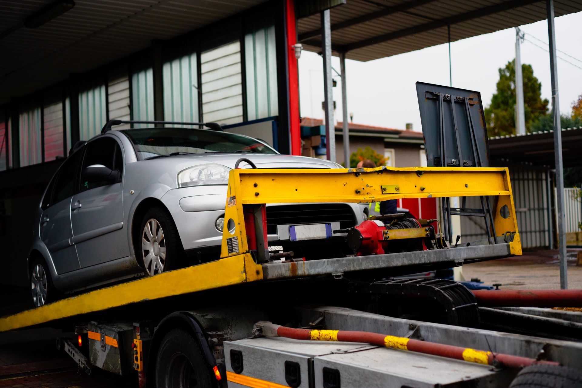 Silver car loaded on a yellow tow truck bed in front of a garage. Silver car loaded on a yellow tow truck bed in front of a garage.