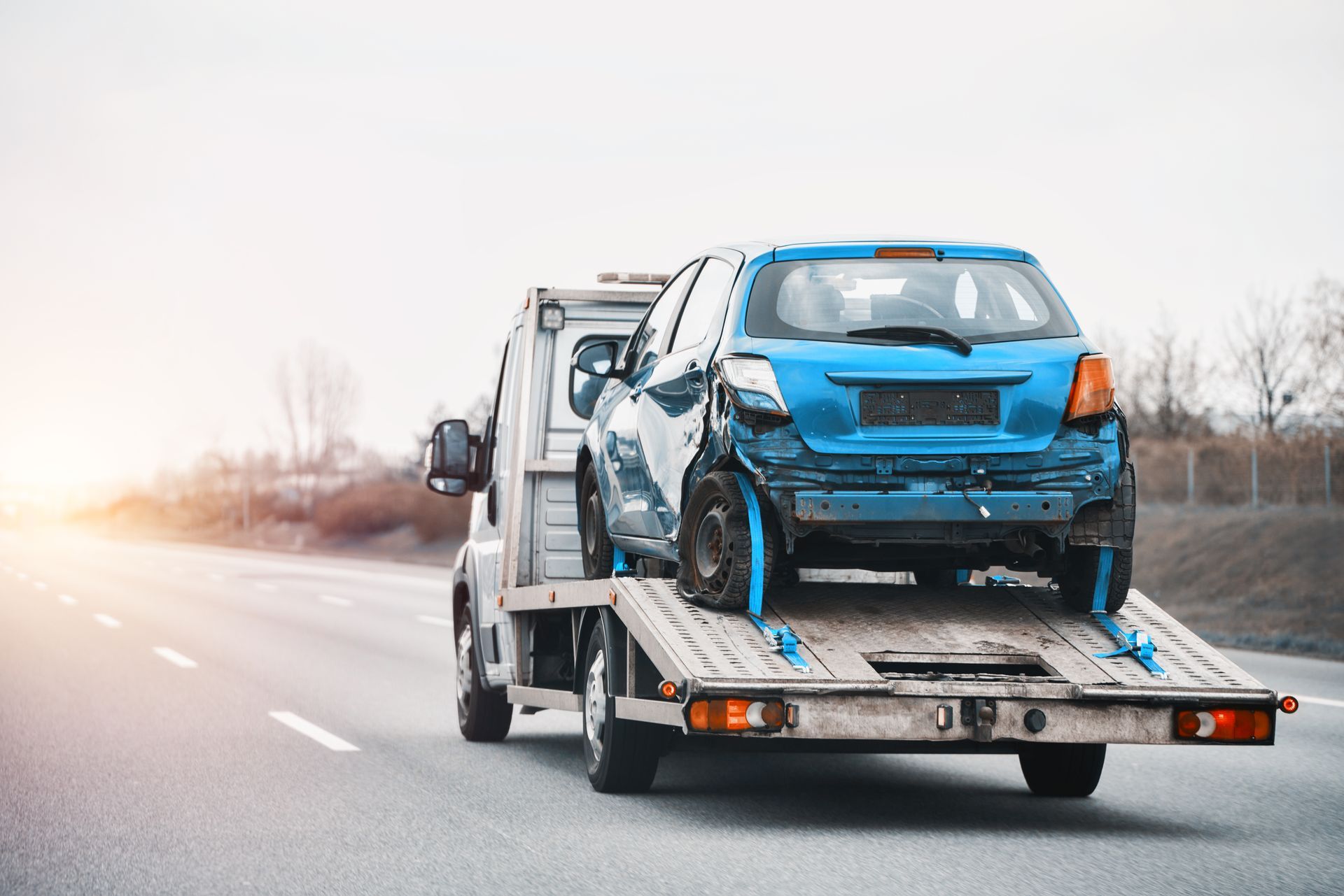Blue, damaged car loaded onto a tow truck on a highway. Blue, damaged car loaded onto a tow truck on a highway.