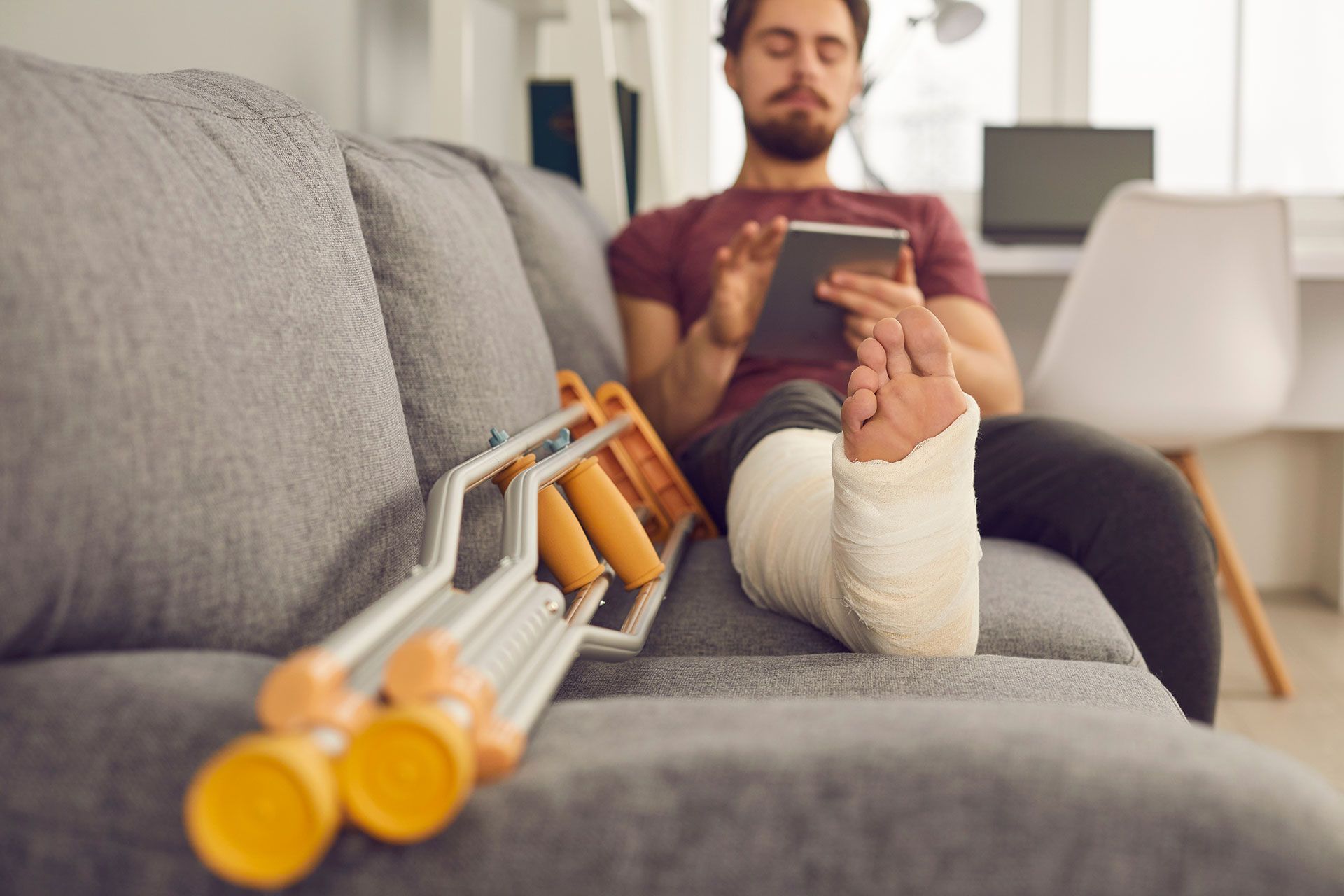 Man with a leg cast, using a tablet while resting on a sofa with crutches nearby.