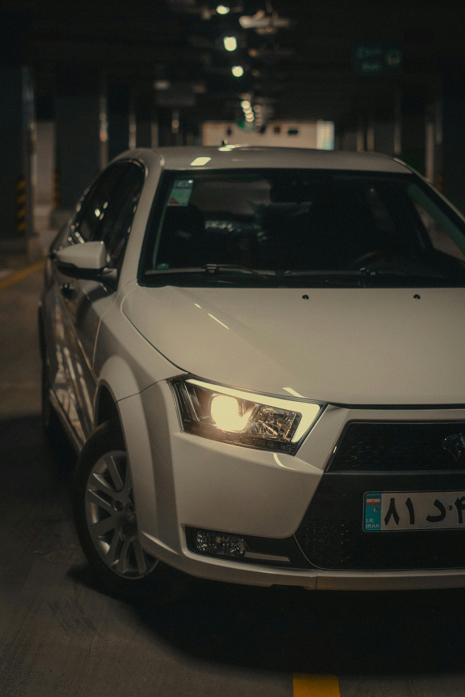 White car parked in a dark garage, headlights on.