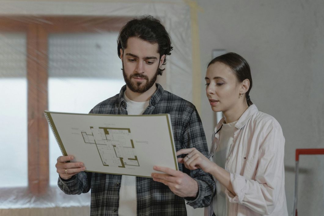 Couple looking at house blueprints in a room under renovation; woman pointing, man holding plans.