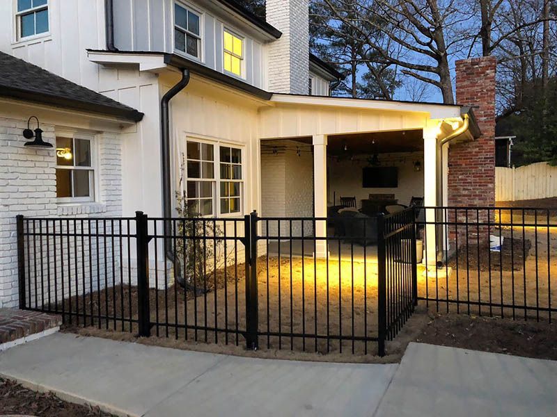 Backyard patio with black metal fence, white house exterior, and a lit covered porch at dusk