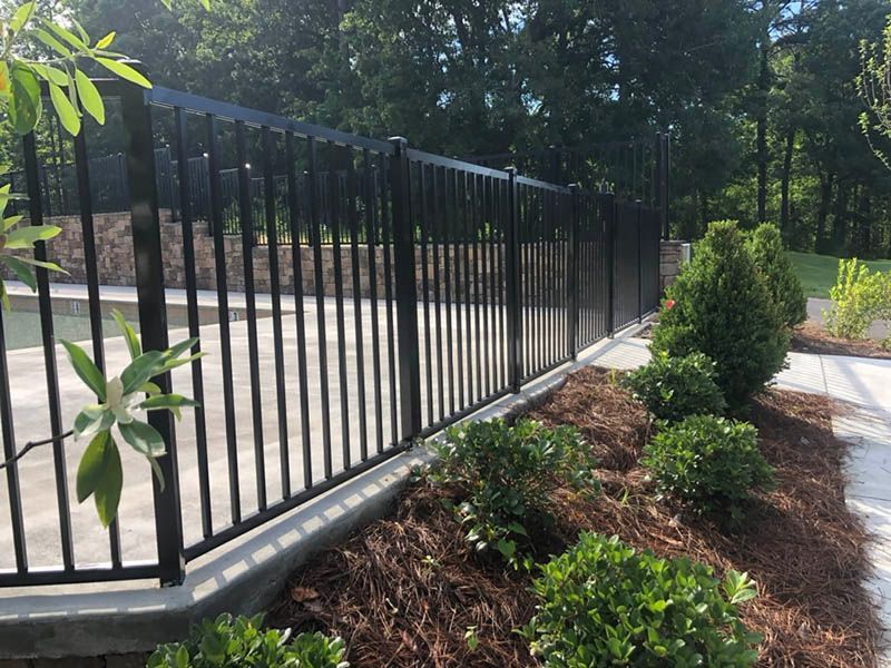 Black metal fence beside mulched landscaping and shrubs along a paved drive