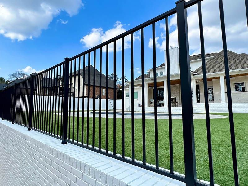 Black metal fence in front of a white brick home with a green lawn and blue sky.