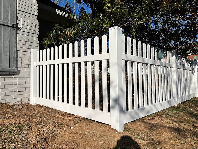 White picket fence enclosing a yard beside a gray house, with dry leaves and grass around it