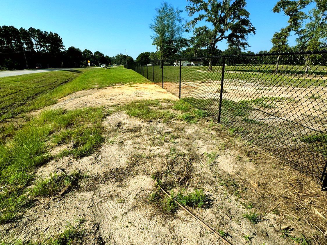 Sandy dirt path beside a chain-link fence and green field under a clear blue sky