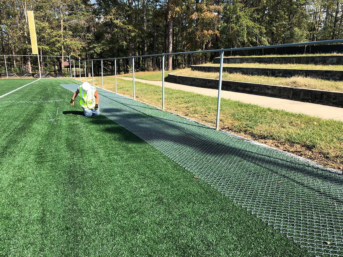 Person in a neon vest kneels by a green sports field fence on a sunny day.