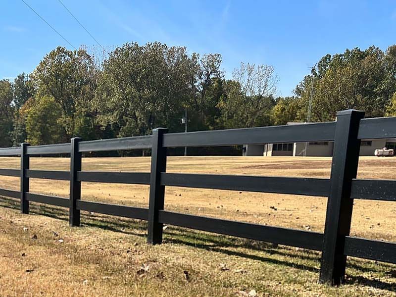 Black wooden fence around a dry field with trees in the background under a clear sky