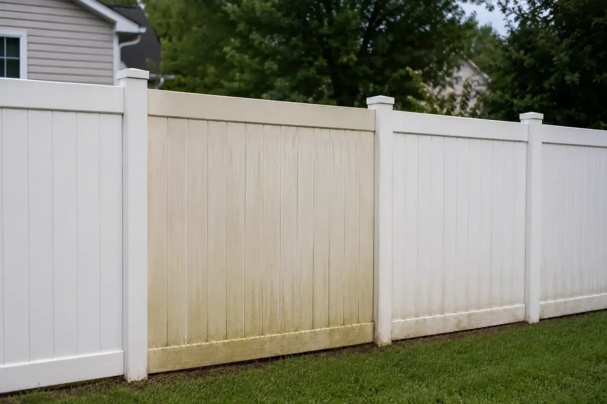 White vinyl fence with one yellowed panel along a grassy yard beside a house
