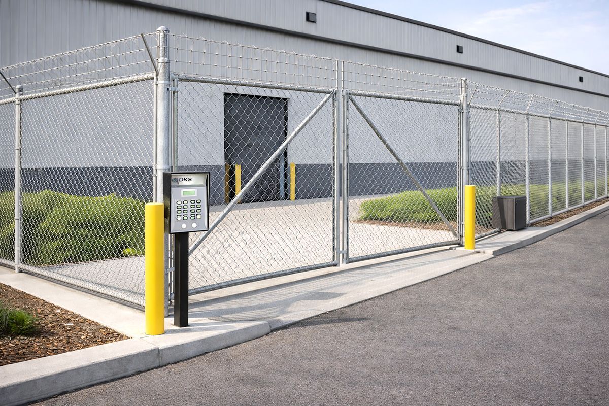 Chain-link security gate beside a gray industrial building with yellow bollards and an intercom keypad