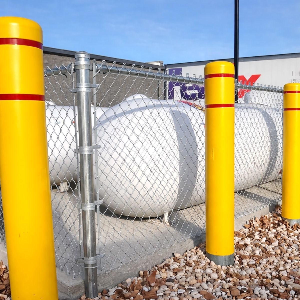 White propane tanks behind a chain-link fence with yellow safety posts and gravel foreground