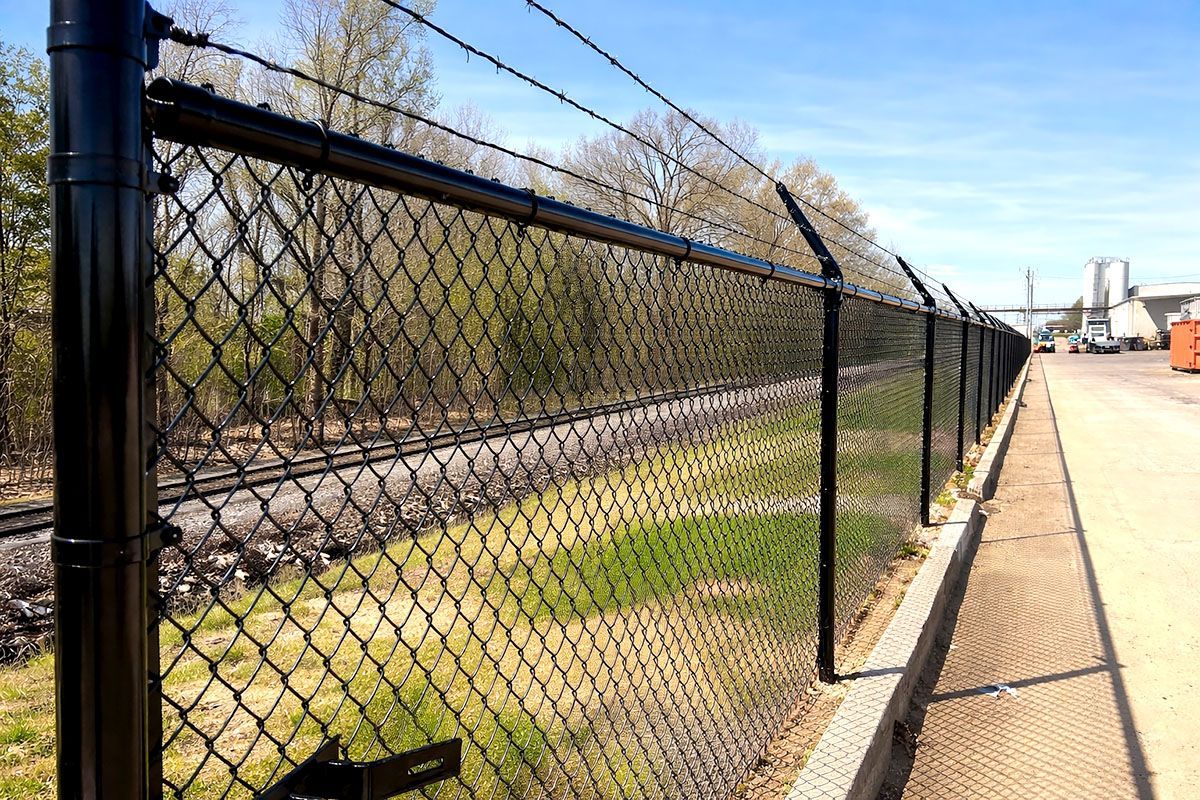 Black chain-link fence beside railroad tracks and a paved walkway under a blue sky.