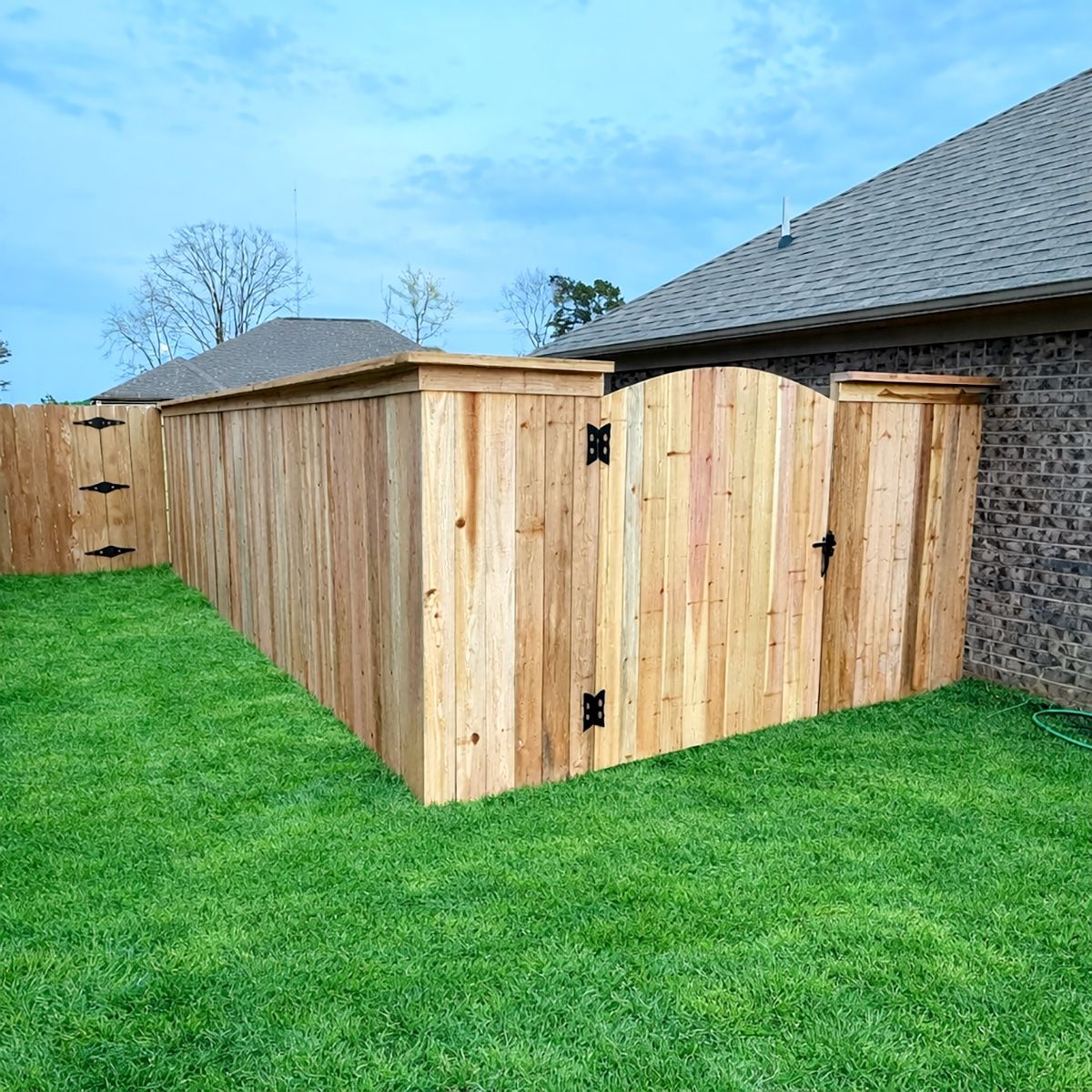 Wooden privacy fence and gate enclosing a backyard beside a brick house on a green lawn