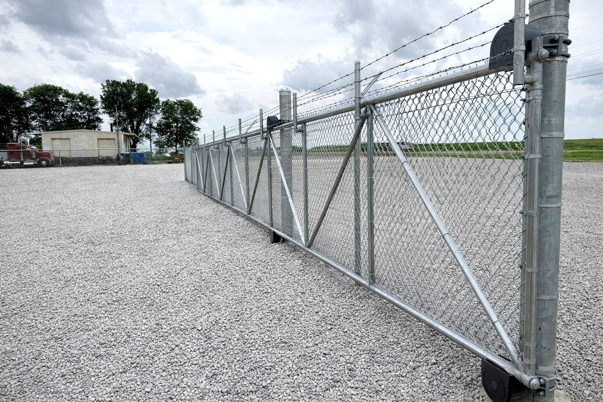 Chain-link sliding gate on a gravel lot beside a fenced industrial area under a cloudy sky