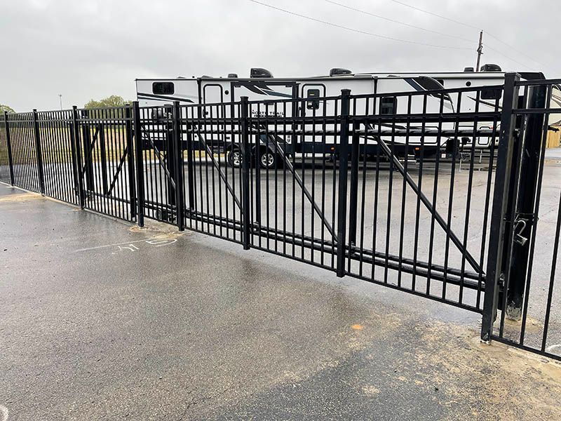 Black metal gate beside a wet pavement, with a parking lot and parked trailer behind it
