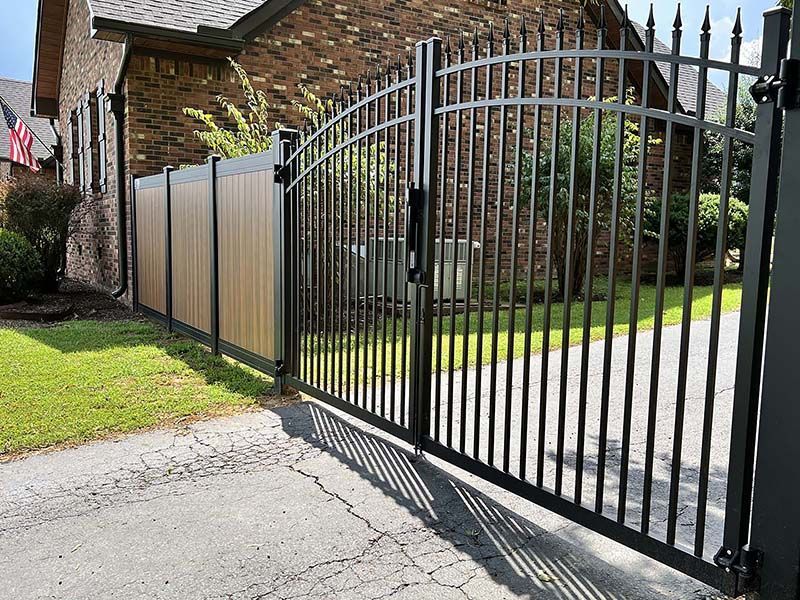 Black metal driveway gate beside a brick house and fenced yard, with a paved driveway in front