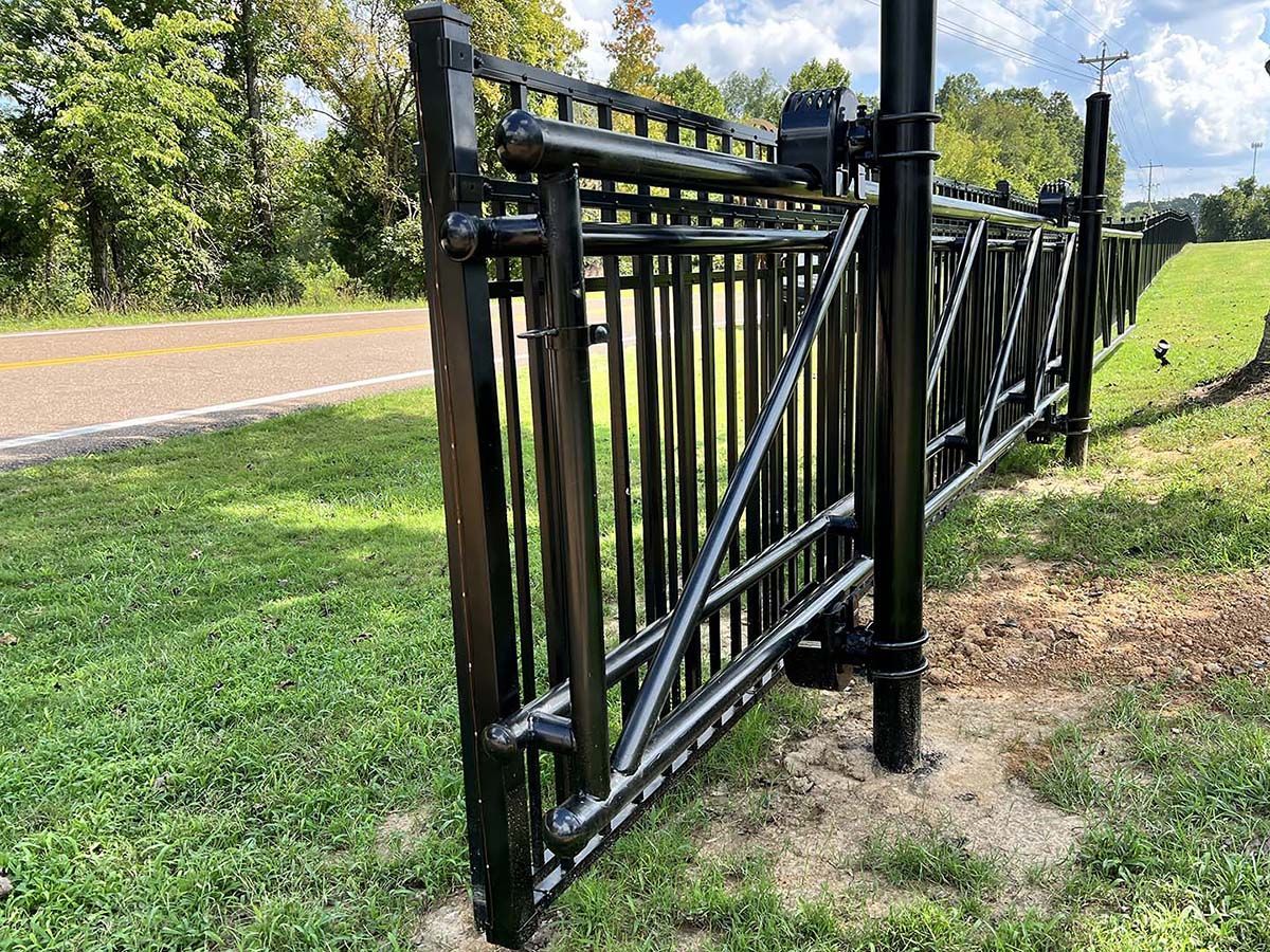 Black metal gate along a roadside, open to a grassy area under bright daylight.