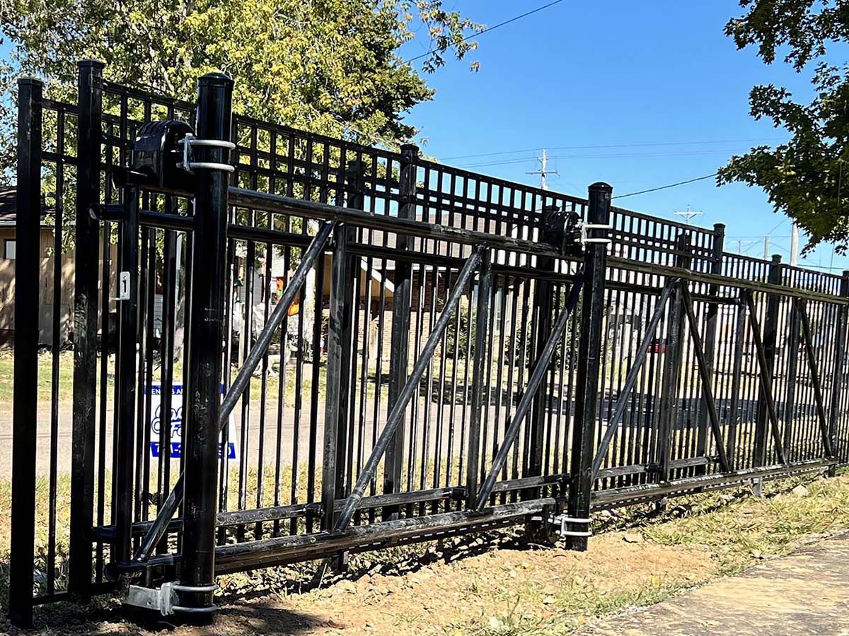 Black metal fence with swinging gate beside a sidewalk, with trees and a lawn behind it.