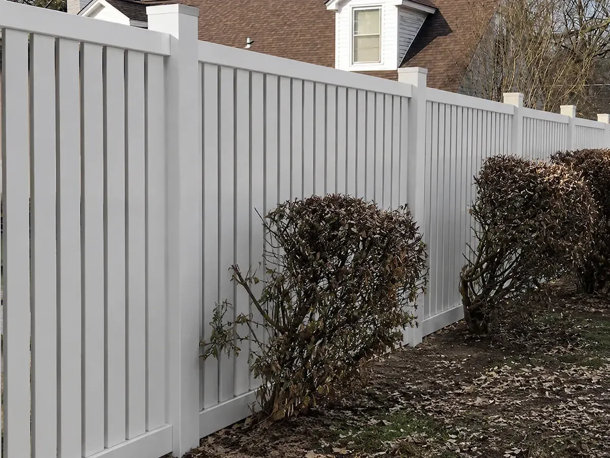 White vinyl fence along a yard with trimmed shrubs and a house in the background