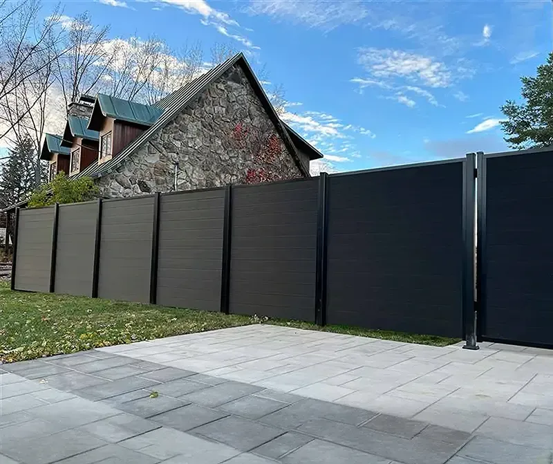 Dark modern fence and gate beside a stone house, with a paved sidewalk and blue sky above.