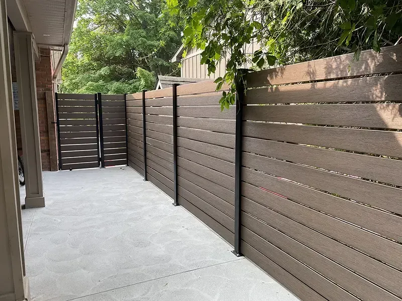Narrow concrete patio walkway bordered by brown horizontal wooden privacy fences and green trees.