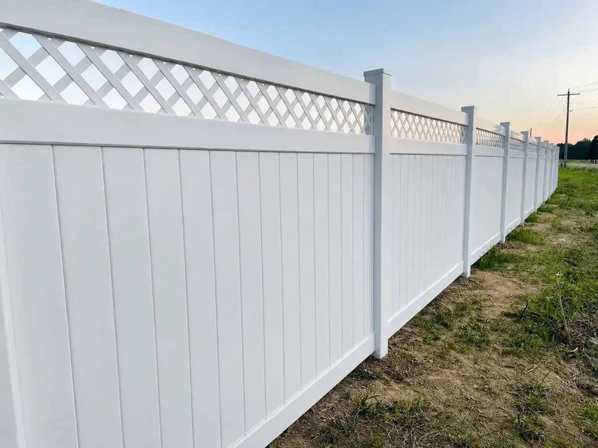 White vinyl privacy fence with lattice top running along a grassy roadside under a clear sky