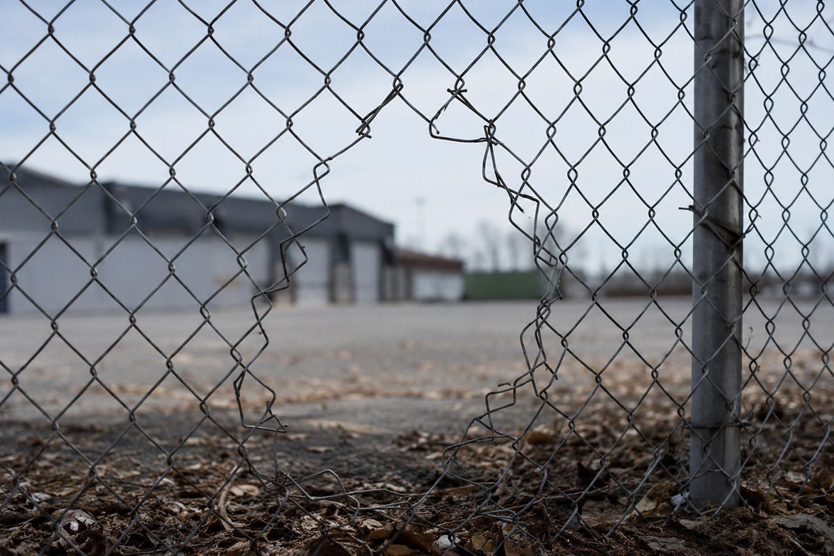 Chain-link fence in foreground with an industrial building and empty lot beyond.