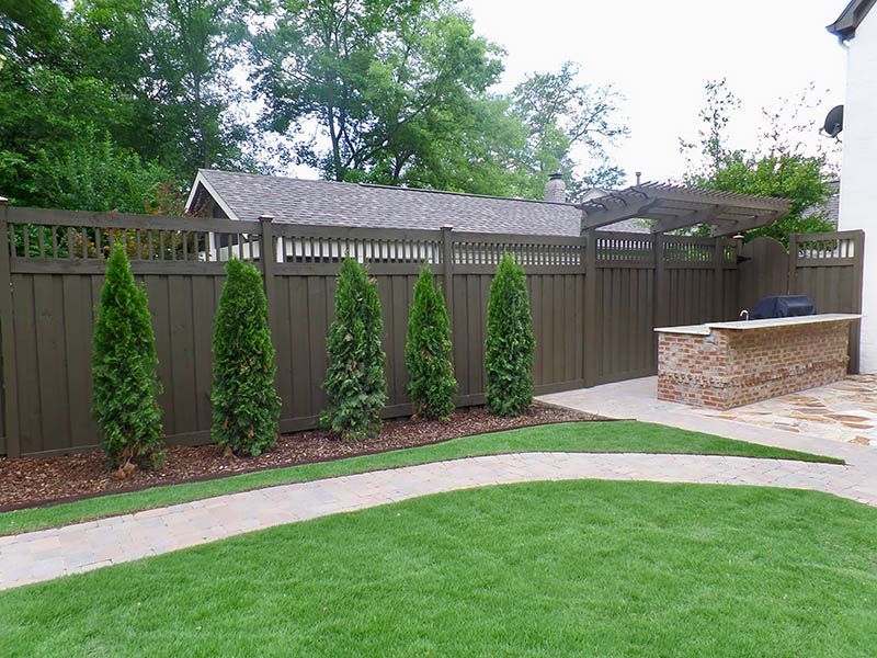 Backyard with green lawn, curved walkway, wooden fence, and small evergreens under trees