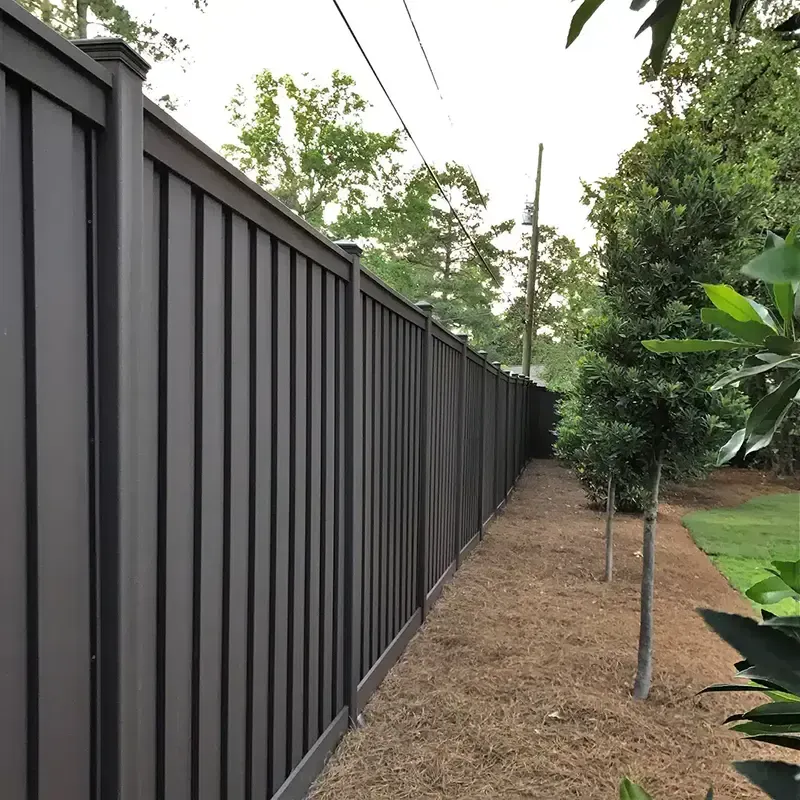 Tall dark metal fence beside a narrow dirt strip with trees and greenery along a property line