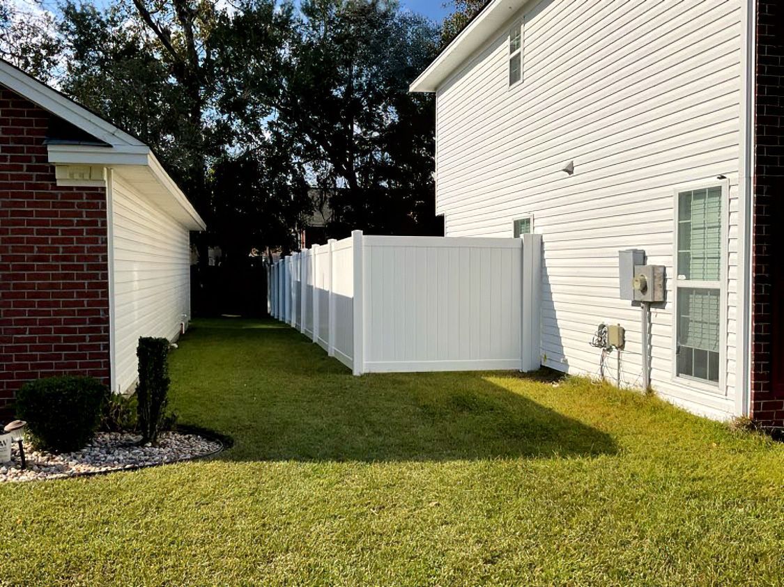 Narrow grassy side yard between brick and white houses, with a white privacy fence and utility meters.