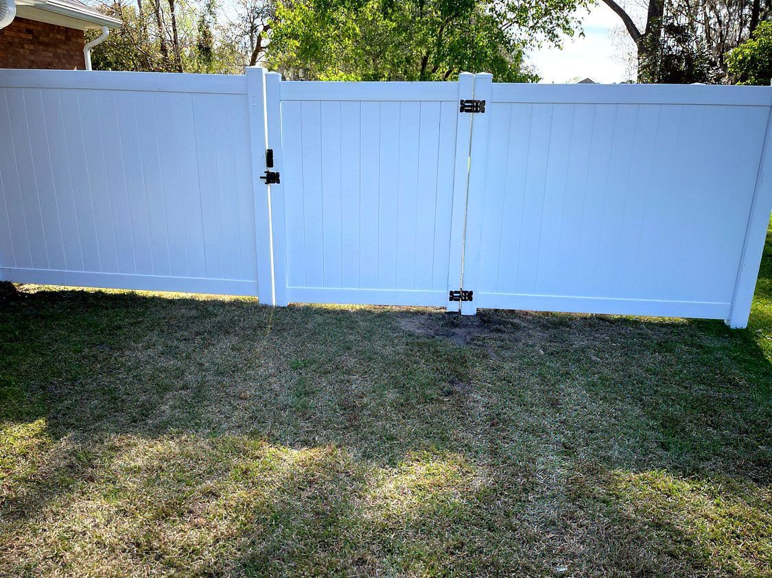 White vinyl fence with a gate in a grassy yard, with trees and sunlight in the background